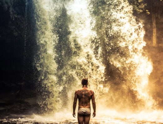 A man is shown standing in water at the base of a waterfall in his underwear