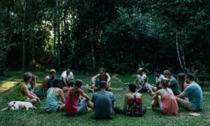 A group of 10 people sit in a circle in the shade on the grass