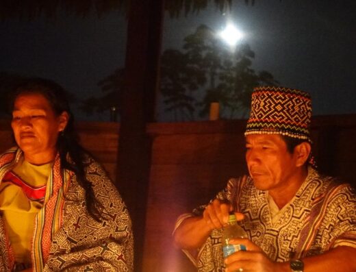 A shaman holds a bottle of medicine before a ceremonial fire, in the light of the moon, while seated next to a shamana