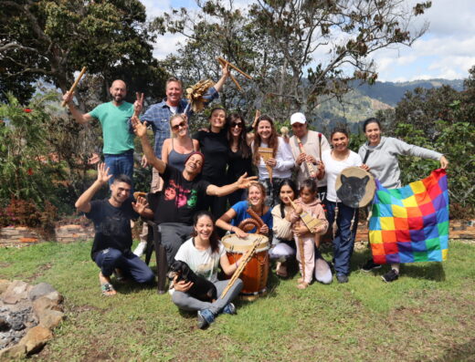 A group of smiling people express joy at the camera while holding musical instruments, a child, a dog, and a colorful flag with mountains and trees in the background
