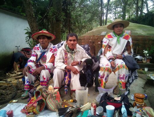 Three men in ceremonial clothing sit in front of what appears to be a container of plant medicine, as well as other candles and other ceremonial items