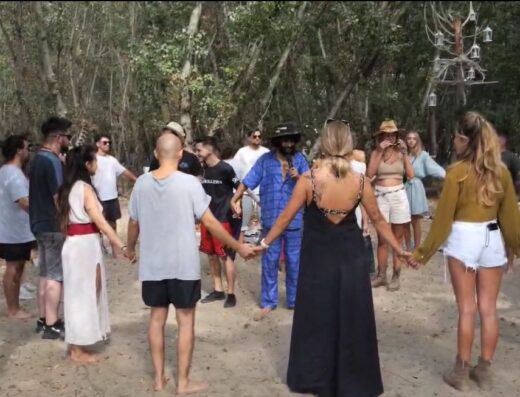A circle of people hold hands in a clearing in the woods