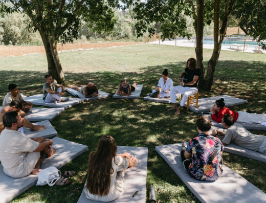 A group of people site in a circle of matts on a large, green, grassy lawn, under the shade of several green, leafy trees, with a Hispanic-looking gentleman playing guitar before an Ayahuasca ceremony